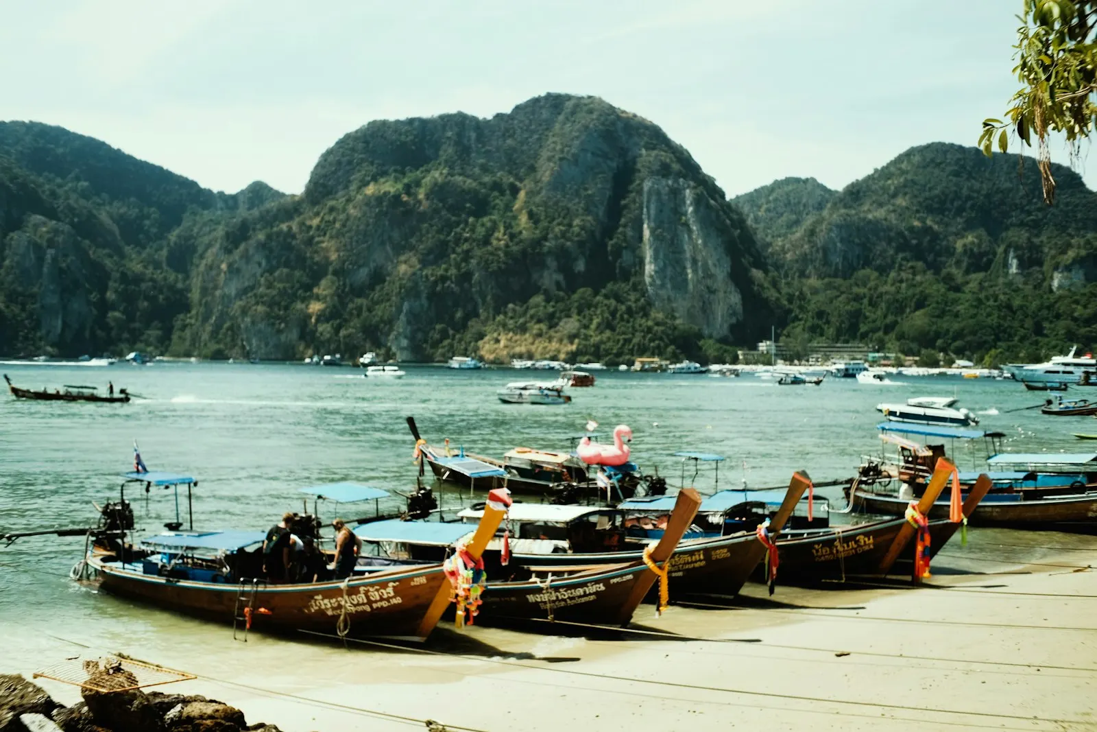 Longtail boats line a sandy beach with mountains behind A serene coastal scene featuring traditional Thai long-tail boats resting on a sunlit sandy beach in the